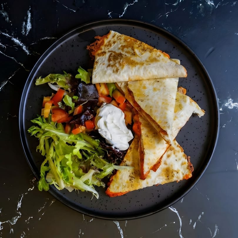 Quesadillas with melted cheese and a side salad with sour cream on a black plate.